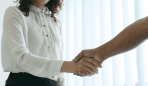 A person in a white long-sleeve shirt and black pants is shaking hands with another person whose arm is visible. The background is bright and softly lit, suggesting a professional or formal setting.