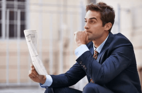 A man in a suit is sitting outdoors, holding a folded newspaper in one hand and resting his chin on his other. He appears to be deep in thought, perhaps pondering the complex taxation of stock options, with a blurred building facade in the background.