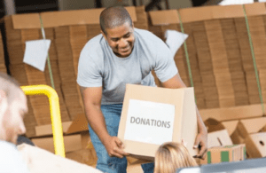 A man is smiling and holding a cardboard box labeled "Donations" while working in a warehouse. Numerous more boxes are stacked in the background. Another person in the foreground is partially visible, also handling boxes for this nonprofit organization, showcasing their investment in community support.