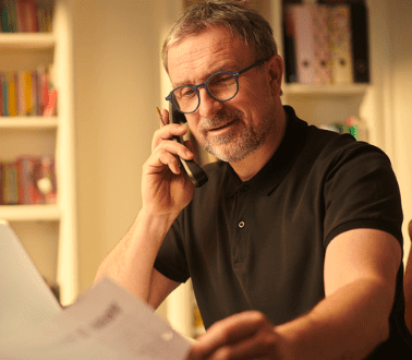A man with short hair and glasses is sitting at a desk, smiling while talking on the phone. He is wearing a black polo shirt and holding a piece of paper. There are bookshelves in the background, suggesting a home or office setting.