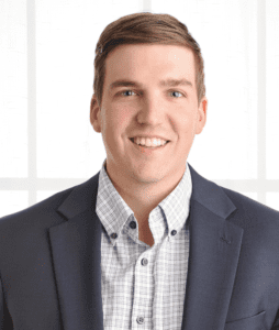 A man with short hair, wearing a dark blazer over a checkered shirt, smiles at the camera. He is standing in front of a light background with a grid pattern.