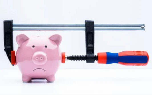 A pink piggy bank with a sad facial expression is being squeezed tightly by a large clamp. The clamp has a red and blue handle, suggesting financial pressure or constraints reminiscent of the recession in 2022. The background is plain white.