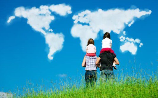 A family of four stands on a grassy hilltop, with two adults carrying two children on their shoulders. They face a clear blue sky where clouds form the shapes of the world map, illustrating the continents and reflecting on global events like economic pain in Russia.