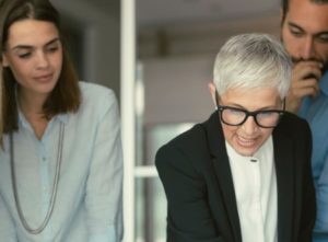 An older woman with short gray hair, a female investor in a black suit, is speaking while two younger colleagues, a woman in a light blue shirt and a man with a beard, listen attentively. They appear to be in a professional setting.