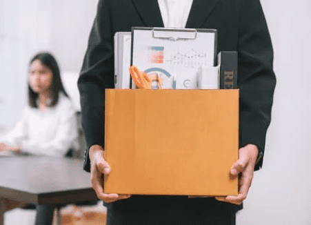 A person in a suit is holding a cardboard box filled with office supplies, including folders, a pen holder, and a few other items. In the background, another person is sitting at a desk. The scene suggests the individual may be exiting corporate life.