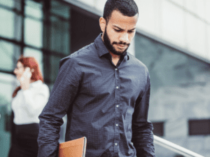 A man in a dark, patterned shirt looks down while walking outside a building, holding a laptop or briefcase. In the background, a woman with red hair is talking on her phone near the building's entrance, possibly discussing succession planning. The scene appears business-oriented.
