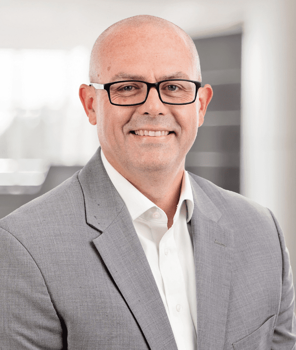 A bald man with glasses is smiling at the camera. He is wearing a grey suit with a white dress shirt. The background is blurred but appears to be an indoor office setting.