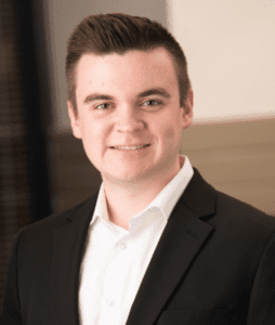 A young man with short brown hair is smiling and wearing a black blazer over a white shirt. He is standing in front of a blurred background with neutral tones.