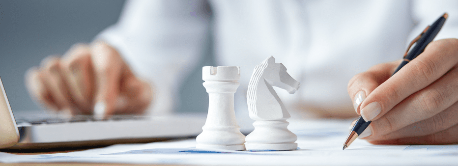 A person working on a laptop with white chess pieces, including a rook and a knight, placed on a document beside them. The person is holding a pen, and their body is slightly out of focus in the background.