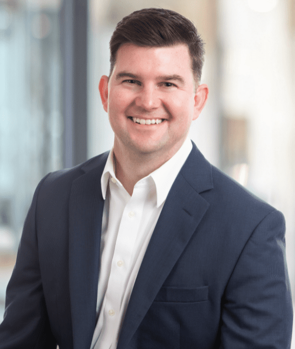 A man with short dark hair is smiling while wearing a navy blue suit jacket and a white dress shirt. He is standing indoors with a blurred background. The overall atmosphere is professional.