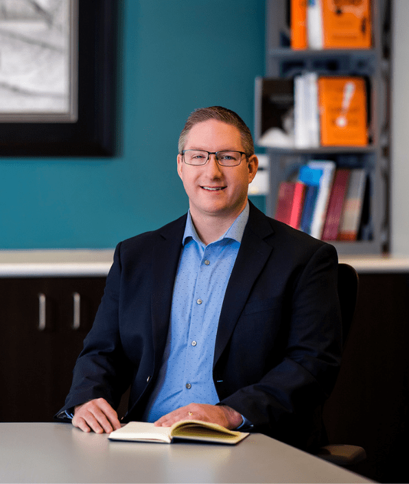 A man wearing a suit jacket and a light blue shirt sits at a table with an open book. He is smiling and looking at the camera. The background features a bookshelf with books and a framed picture on the wall.