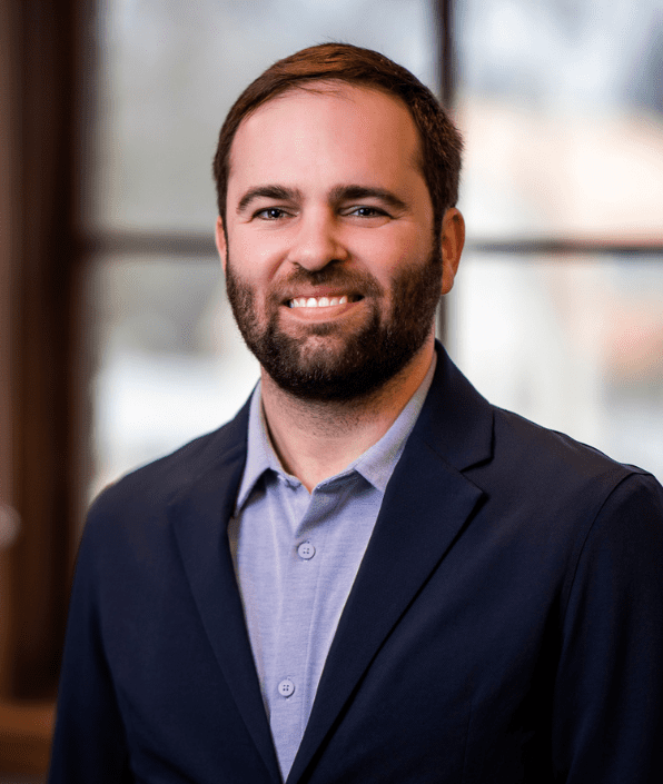 A man with short brown hair and a beard is smiling at the camera. He is wearing a blue button-up shirt and a dark blazer. The background is softly blurred, suggesting an indoor setting near a window.