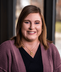 A person with shoulder-length brown hair, wearing a purple cardigan and a black top, smiles at the camera. They have a gold necklace and are in front of a window, with blurred outdoor scenery in the background.