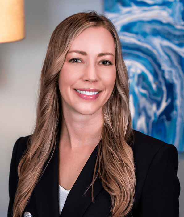 A woman with long, light brown hair smiles while wearing a dark blazer over a white top. She is standing in an indoor setting with a blue and white abstract painting in the background.