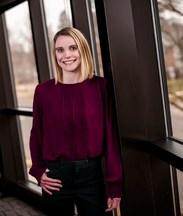 A woman with shoulder-length blonde hair, wearing a maroon blouse and black pants, stands inside a building near tall windows. She is smiling and has one hand in her pocket. Outside the windows, a tree and parts of a road are visible in the blurred background.