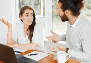 A woman and a man are having a discussion at a table. The woman gestures with her right hand raised and appears to be explaining something about financial planning. The man also gestures with his hands. They have papers and a laptop in front of them. The setting looks like an office.