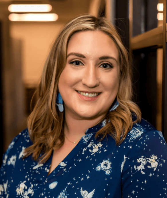A woman with light brown hair smiles while standing indoors. She is wearing a dark blue blouse with various small, white designs and blue earrings. The background is softly lit with what appears to be a hallway or interior space.