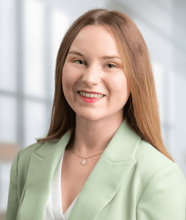 A woman with straight, light brown hair is smiling. She is dressed in a light green blazer over a white top and is wearing a necklace with a pendant. The background features out-of-focus large windows, indicating an indoor setting with natural light.