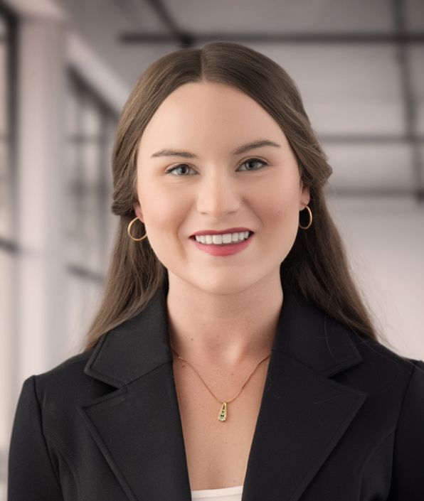 A woman with straight, light brown hair is smiling. She is dressed in a black blazer over a white top and is wearing a necklace with a pendant. The background features out-of-focus large windows, indicating an indoor setting with natural light.