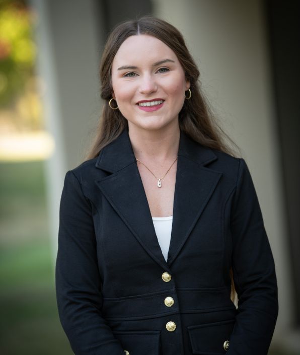 A woman with straight, light brown hair is smiling. She is dressed in a black blazer over a white top and is wearing a necklace with a pendant. The background features out-of-focus outdoors, green with a white building pillar.