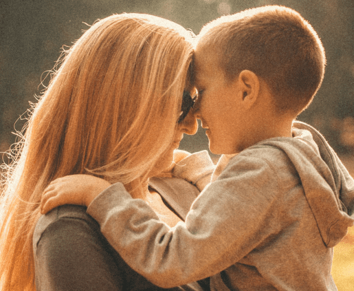 A woman with long blonde hair and sunglasses embraces a young boy wearing a hooded sweatshirt. They are forehead to forehead, sharing a tender moment outdoors, with soft sunlight illuminating them. The background is blurred.