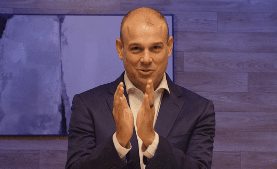 A man wearing a navy blue suit and white shirt stands in front of a blurred background with a television screen. He is gesturing with his hands close together, appearing to be in mid-speech.