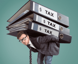 A man in a suit, glasses, and a striped tie bends under the heavy weight of three large binders labeled "TAX" stacked on his back. He appears strained as he balances the oversized folders, symbolizing the burden of estate tax mitigation, against a green background.