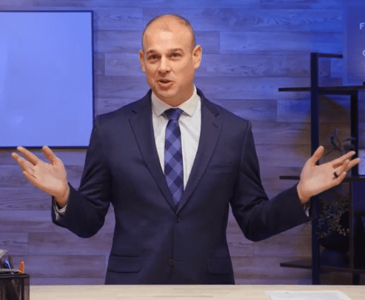 A man in a navy suit and blue tie stands behind a desk, gesturing with both hands. The background features a wooden wall, a whiteboard on the left, and a shelving unit on the right with books and decor items. The scene appears to be in an office or studio setting, likely discussing the volatile market conditions.