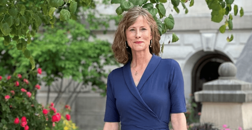 A woman with shoulder-length brown hair smiles while standing outside in front of a building. She is wearing a navy blue dress and there are green trees and pink flowers in the background.