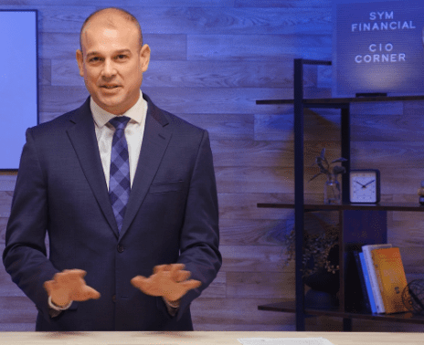 A man in a navy suit and blue checkered tie is speaking and gesturing with his hands. He stands behind a desk with a sleek, minimalist background, including shelves with books, a clock, and a plant. A sign reads "SYM Financial CIO Corner" in the top right.