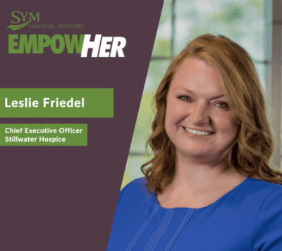 Leslie Friedel, Chief Executive Officer of Stillwater Hospice, smiles in a blue top. The image features the SYM Financial Advisors logo and a green banner with the word "EmpowHER," highlighting her commitment to palliative care.