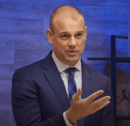 A bald man in a blue suit and tie is speaking animatedly, gesturing with his right hand. He is standing in front of a wooden textured wall with a blue tone, discussing artificial intelligence. A black shelf can be seen in the background.