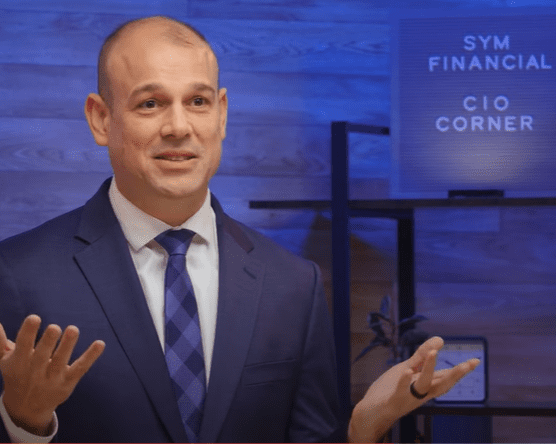 A bald man in a blue suit and checkered tie stands gesturing with both hands. Behind him, a sign reads "SYM Financial CIO Corner." The background includes a blue-lit wooden wall and a standing shelf, which features a book on artificial intelligence. He appears to be engaged in conversation or presenting.
