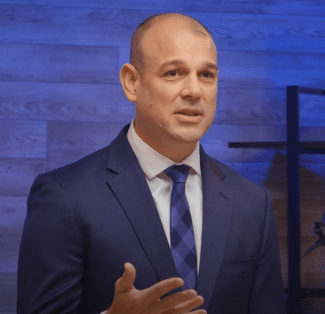 A man in a blue suit with a white shirt and blue checkered tie is speaking. Gesturing with his right hand, he stands against a wood-paneled wall accented by blue lighting and a black shelf. Discussing artificial intelligence, his words resonate within the modern setting.