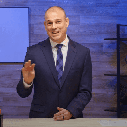 A bald man in a blue suit with a patterned tie stands at a wooden desk, gesturing with his right hand. He is smiling and appears to be discussing artificial intelligence. The background features a wooden wall with blue lighting, a screen, and shelving.