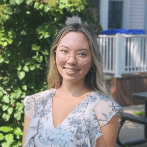 A person with long, blonde hair and glasses smiles outdoors. They are wearing a light-colored, floral-patterned blouse. The background features green foliage and part of a house with white railings and a window.