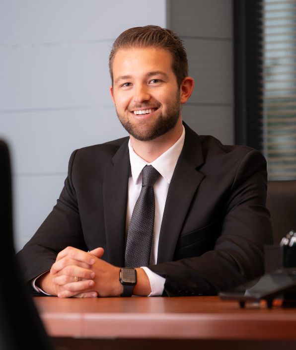 young man with brown hair and a beard in suit and tie sitting at a desk
