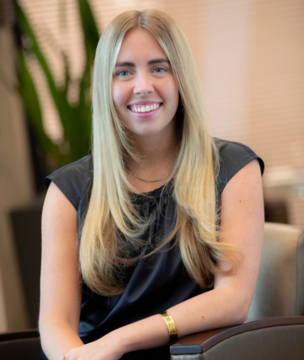 young woman with long blond hair in a black blouse sitting on a chair leaning in and smiling