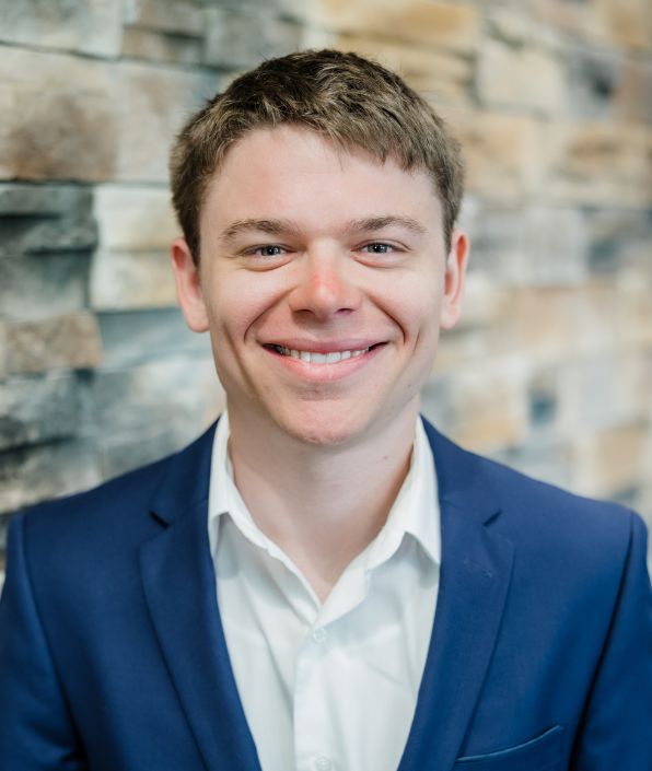 top half of young man standing in front of a brick wall with short brown hair in a blue suit coat
