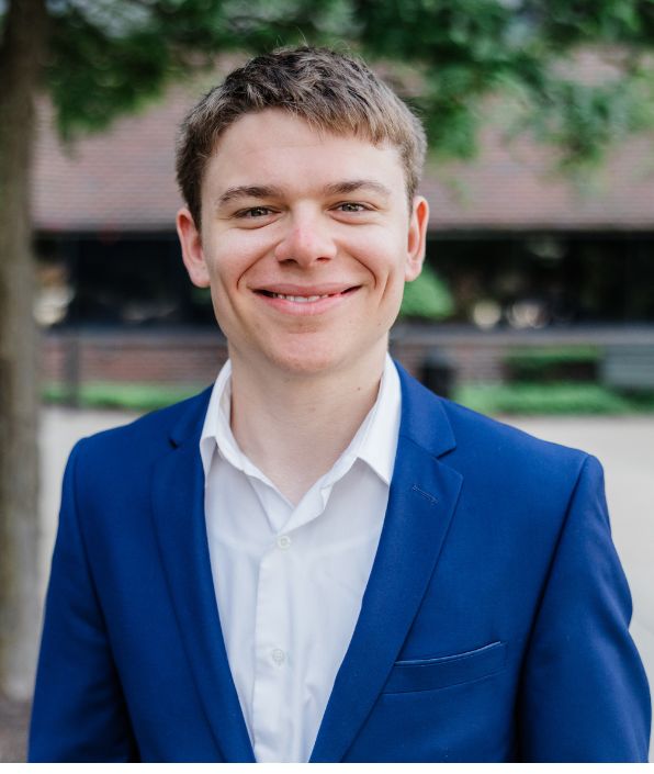 top half of young man standing outside of an office building with short brown hair in a blue suit coat