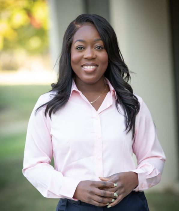 woman with black long hair and dark skin standing outside in a pink button down shirt with her hands folded in a relaxed manner.