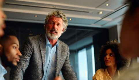 a man with gray hair and beard in a tweed suitcoat speaks among other professionals in a meeting room