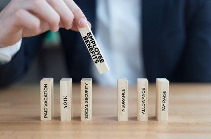 A hand is holding a wooden block with the label "employee benefits" listed on it. The is a stack on the table of other blocks set up like dominoes