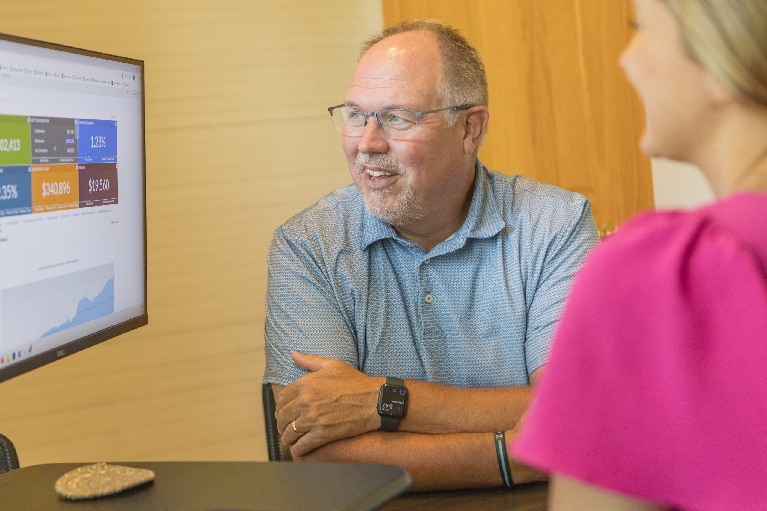 a middle aged man in a blue golf shirt with glasses reviewing a portfolio report on a computer screen with a woman in a pink dress