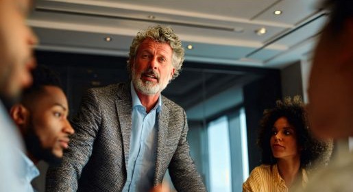 A man with a beard in a tweed jacket works around a board table with fellow employees