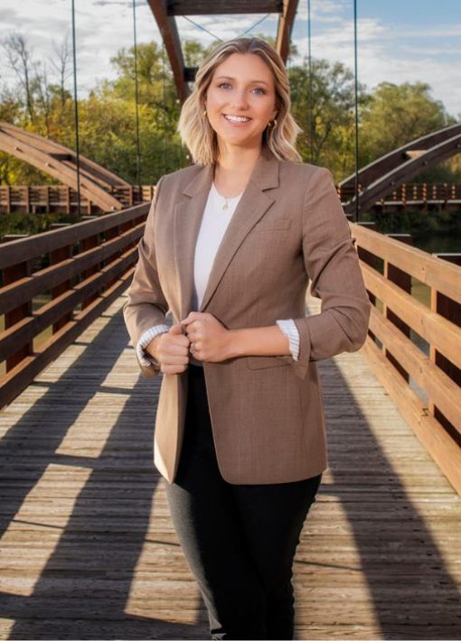 standing on a bridge is a woman with blond hair and blue eyes in a tan blazer with white undershirt