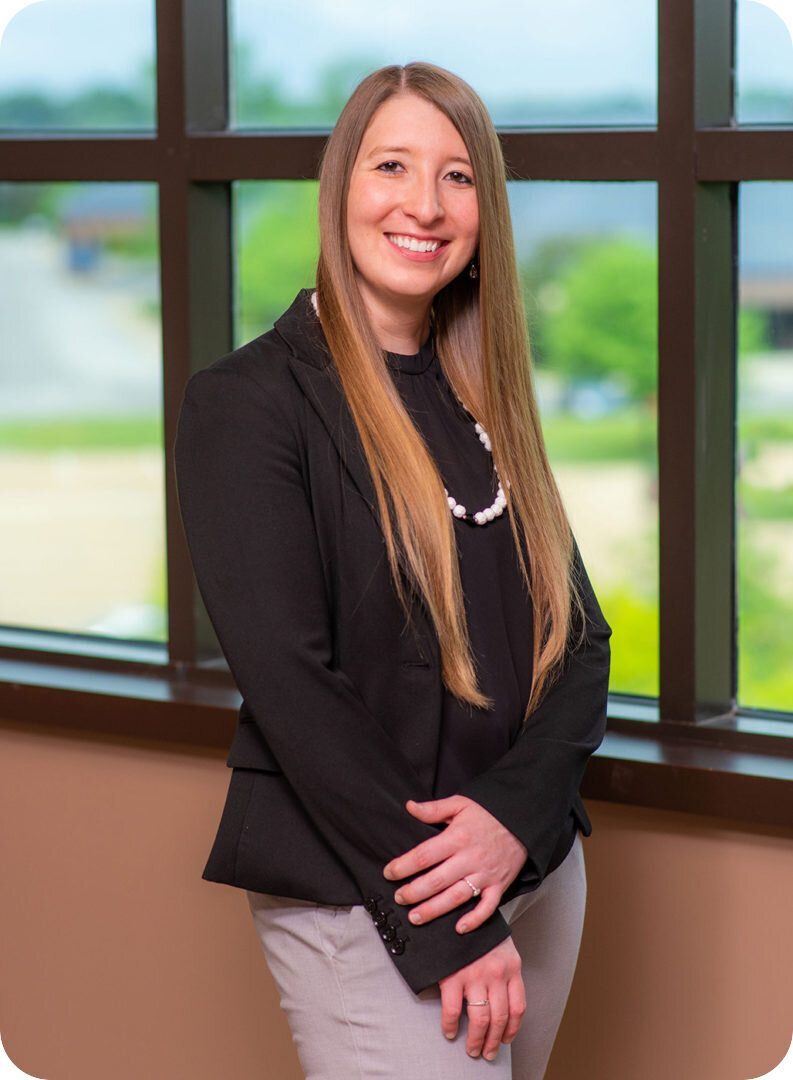 rsz_2team-individual-secondary A woman with long, straight brown hair wearing a black blazer, black blouse, and light gray pants stands in front of a large window. She is smiling and has her hands clasped in front of her. The window shows a blurred outdoor background.